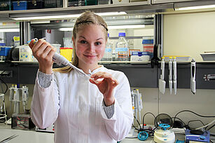 Ein junges Mädchen guckt in die Kamera und pipettiert dabei Flüssigkeit in ein Röhrchen. Im Hintergrund: Laborumfeld.